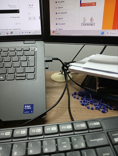 A cluttered desk setup with two monitors, a laptop displaying software code, a keyboard, some papers, and scattered small blue electronic components. (Captioned by AI)